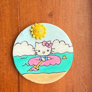 Op de afbeelding: Een handgeschilderde keramische onderzetter met Hello Kitty in een roze binnenband die op turquoise water drijft met een gele zon en witte wolken.