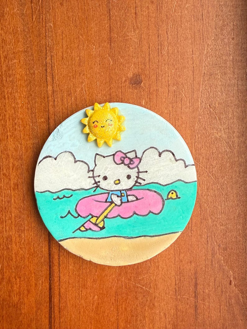 Op de afbeelding: Een handgeschilderde keramische onderzetter met Hello Kitty in een roze binnenband die op turquoise water drijft met een gele zon en witte wolken.