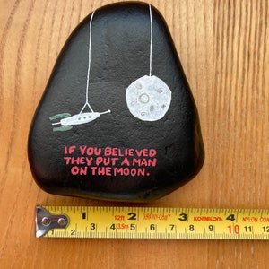 May include: A black painted rock with a white moon and a spaceship hanging from strings. The rock has the text "IF YOU BELIEVED THEY PUT A MAN ON THE MOON." written in pink.