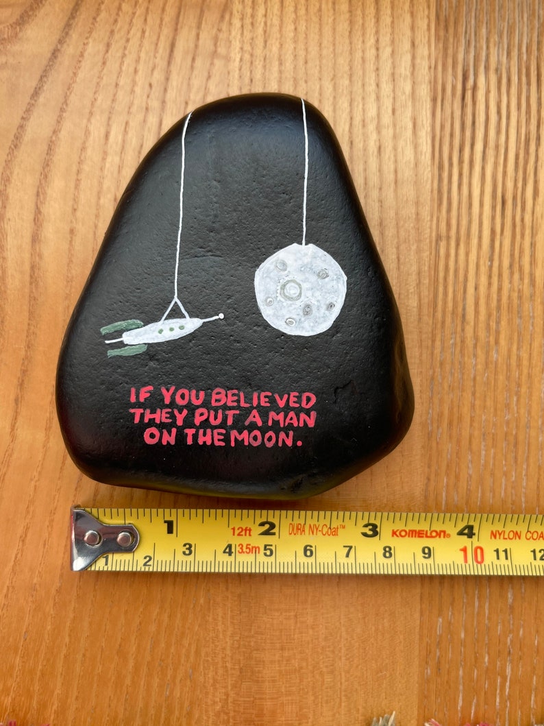 May include: A black painted rock with a white moon and a spaceship hanging from strings. The rock has the text "IF YOU BELIEVED THEY PUT A MAN ON THE MOON." written in pink.
