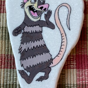 May include: Hand-painted rock art of a cartoon opossum with a pink nose and tail, black and gray stripes, and a white background.