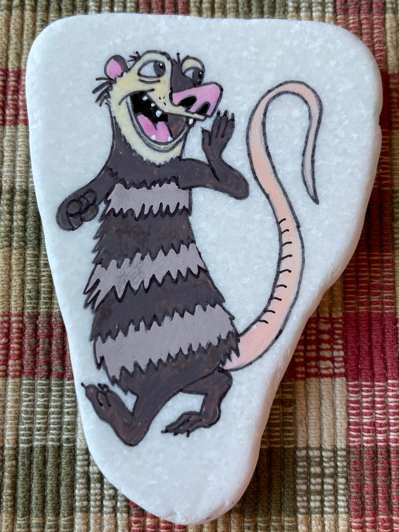 May include: Hand-painted rock art of a cartoon opossum with a pink nose and tail, black and gray stripes, and a white background.