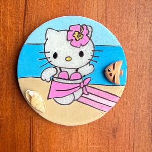 Op de afbeelding: Een ronde keramische onderzetter met een geschilderd beeld van Hello Kitty, gekleed in een roze badpak en een roze bloem op haar hoofd. De onderzetter is geplaatst tegen een blauwe oceaanachtergrond met een wit zandstrand en een kleine oranje vis.