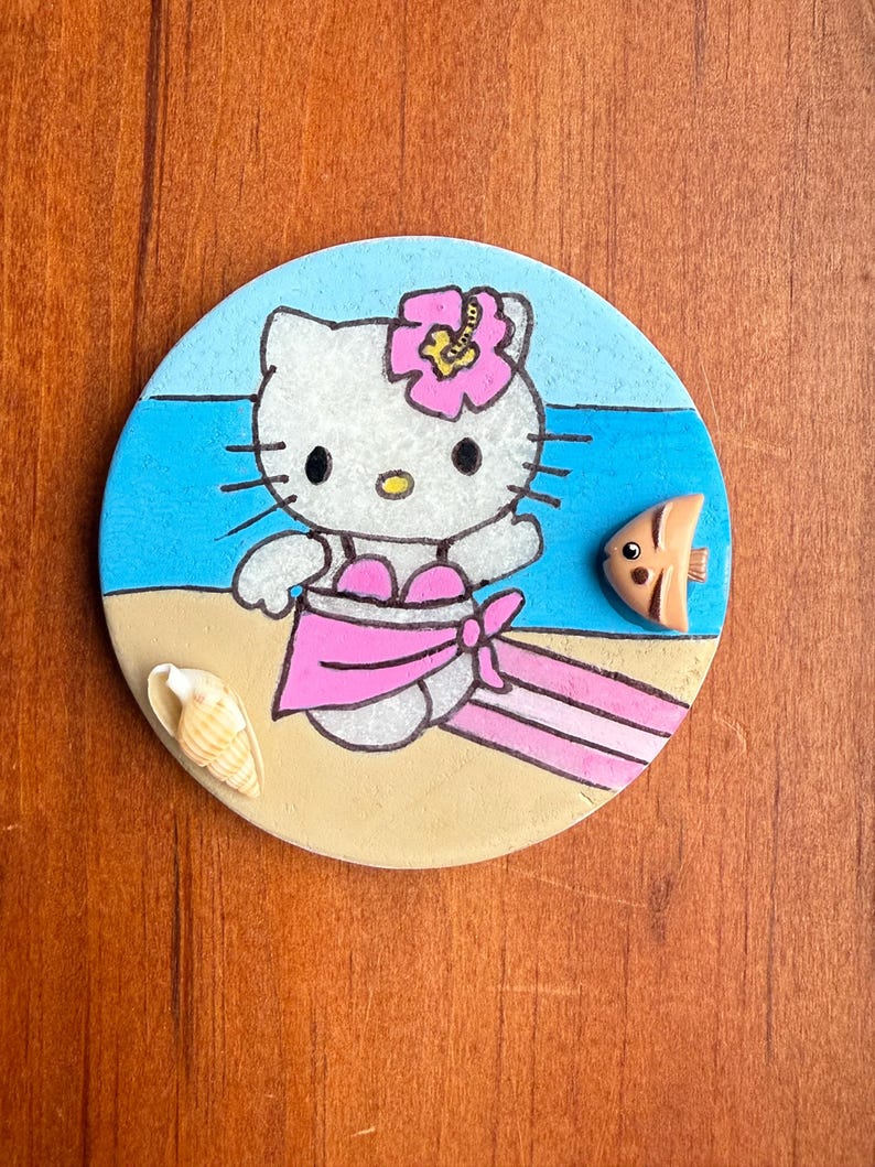 Op de afbeelding: Een ronde keramische onderzetter met een geschilderd beeld van Hello Kitty, gekleed in een roze badpak en een roze bloem op haar hoofd. De onderzetter is geplaatst tegen een blauwe oceaanachtergrond met een wit zandstrand en een kleine oranje vis.