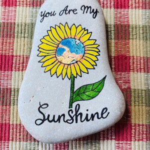 Puede incluir: Una piedra pintada de gris con un girasol amarillo pintado en ella. El girasol tiene un centro azul y naranja. Las palabras "You are my Sunshine" est&aacute;n escritas en pintura negra en la piedra.