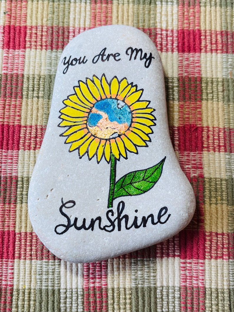 Puede incluir: Una piedra pintada de gris con un girasol amarillo pintado en ella. El girasol tiene un centro azul y naranja. Las palabras "You are my Sunshine" est&aacute;n escritas en pintura negra en la piedra.