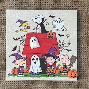 May include: A square decorative tile featuring a Peanuts Halloween illustration with Snoopy, Charlie Brown, and Lucy. The scene includes a red doghouse, pumpkins, ghosts, and bats. A small 3D white ghost and an orange pumpkin are attached to the surface.