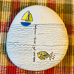 May include: Hand-painted rock with a yellow and blue sailboat on top of wavy blue lines. The text "holy cow I think I got one here" is written vertically on the right side of the rock. A yellow fish with black stripes is swimming in the water.