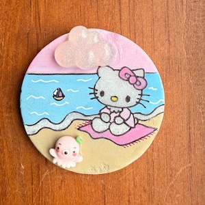 Op de afbeelding: Een ronde, beschilderde keramische onderzetter met Hello Kitty die op een roze handdoek op het strand zit. Een roze octopus met een groen blaadje bevindt zich in het zand. Een witte wolk met glitter bevindt zich in de linkerbovenhoek.