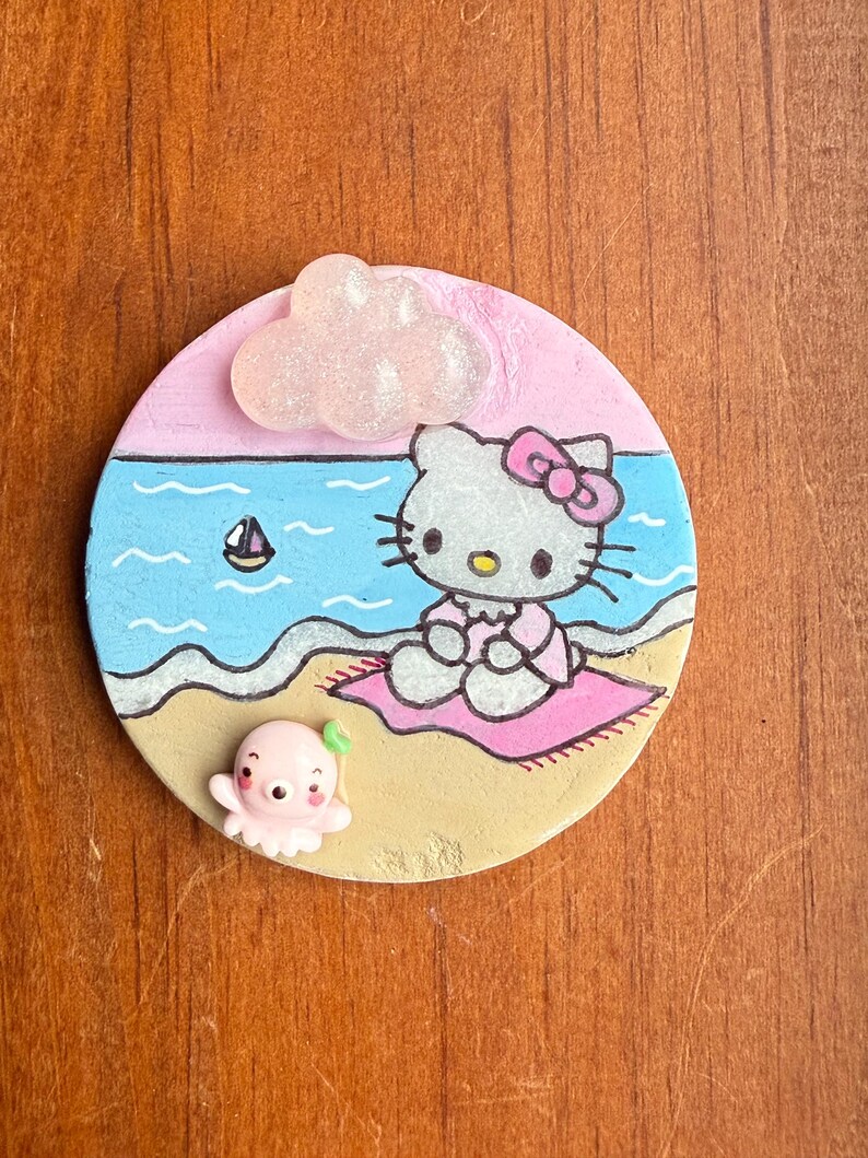 Op de afbeelding: Een ronde, beschilderde keramische onderzetter met Hello Kitty die op een roze handdoek op het strand zit. Een roze octopus met een groen blaadje bevindt zich in het zand. Een witte wolk met glitter bevindt zich in de linkerbovenhoek.