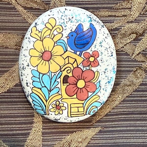 May include: Oval decorative stone with a painted floral and bird design. The design features yellow and red flowers, blue leaves, and a blue bird perched on a yellow birdhouse. The background is white with blue glitter accents.