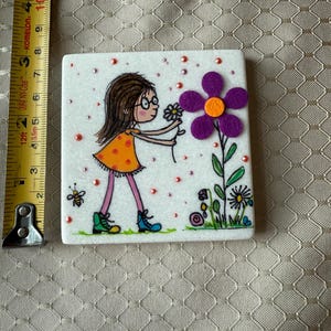 Puede incluir: Azulejo de cer&aacute;mica cuadrado con una ilustraci&oacute;n dibujada a mano de una ni&ntilde;a con un vestido naranja recogiendo una flor. El azulejo tiene un fondo blanco con detalles en rosa y naranja. Tambi&eacute;n se representa una abeja y otras flores. El azulejo mide aproximadamente 7.6 cm.