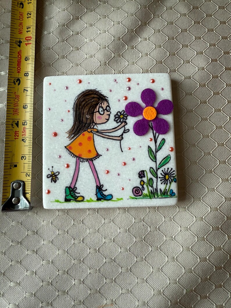 Puede incluir: Azulejo de cer&aacute;mica cuadrado con una ilustraci&oacute;n dibujada a mano de una ni&ntilde;a con un vestido naranja recogiendo una flor. El azulejo tiene un fondo blanco con detalles en rosa y naranja. Tambi&eacute;n se representa una abeja y otras flores. El azulejo mide aproximadamente 7.6 cm.