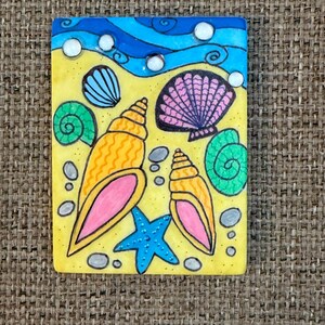 May include: A rectangular, hand-painted artwork depicting a beach scene. The artwork features a yellow sandy area with seashells, a blue starfish, and a blue ocean with white bubbles. The seashells are in shades of orange, pink, green, and blue.