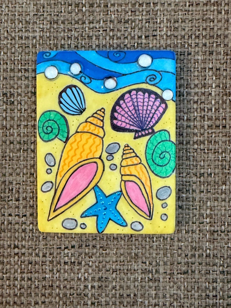 May include: A rectangular, hand-painted artwork depicting a beach scene. The artwork features a yellow sandy area with seashells, a blue starfish, and a blue ocean with white bubbles. The seashells are in shades of orange, pink, green, and blue.