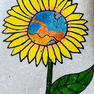 Puede incluir: Una piedra pintada a mano con un girasol amarillo con un globo terr&aacute;queo azul, naranja y gris en el centro. El girasol tiene un tallo y una hoja verdes.