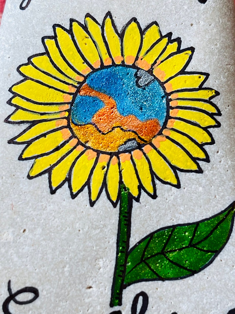 Puede incluir: Una piedra pintada a mano con un girasol amarillo con un globo terr&aacute;queo azul, naranja y gris en el centro. El girasol tiene un tallo y una hoja verdes.