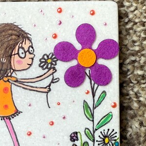 Puede incluir: Una ilustraci&oacute;n caprichosa presenta a una ni&ntilde;a con un vestido naranja y zapatos azules ofreciendo una flor a una gran flor de fieltro morado con un centro naranja. El fondo es blanco con peque&ntilde;os puntos rosas y naranjas y otras flores dibujadas.