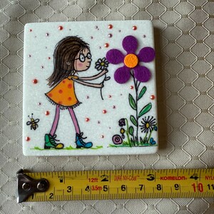 Puede incluir: Azulejo de cer&aacute;mica blanco cuadrado con una ilustraci&oacute;n caprichosa. Una ni&ntilde;a con un vestido naranja y gafas se estira hacia una gran flor morada. El azulejo est&aacute; decorado con peque&ntilde;as cuentas rojas y una abeja.