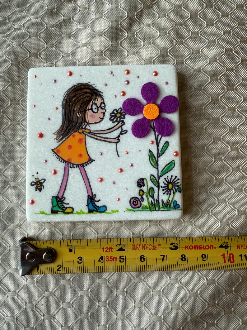 Puede incluir: Azulejo de cer&aacute;mica blanco cuadrado con una ilustraci&oacute;n caprichosa. Una ni&ntilde;a con un vestido naranja y gafas se estira hacia una gran flor morada. El azulejo est&aacute; decorado con peque&ntilde;as cuentas rojas y una abeja.
