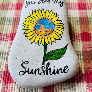 Puede incluir: Piedra pintada con un dise&ntilde;o de girasol y las palabras "You are my Sunshine" en cursiva negra.