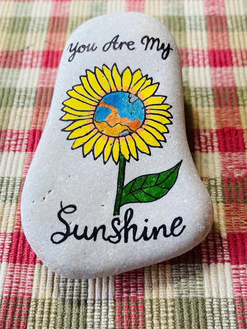 Puede incluir: Piedra pintada con un dise&ntilde;o de girasol y las palabras "You are my Sunshine" en cursiva negra.