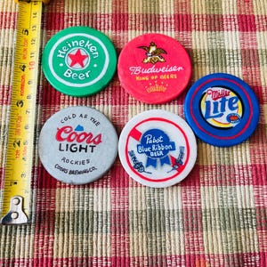 Puede incluir: Cinco fichas de cerveza redondas y de colores con diferentes logotipos de marcas. Las fichas son verde, roja, blanca, azul y morada. Los logotipos incluyen Heineken, Budweiser, Coors Light, Pabst Blue Ribbon y Miller Lite.