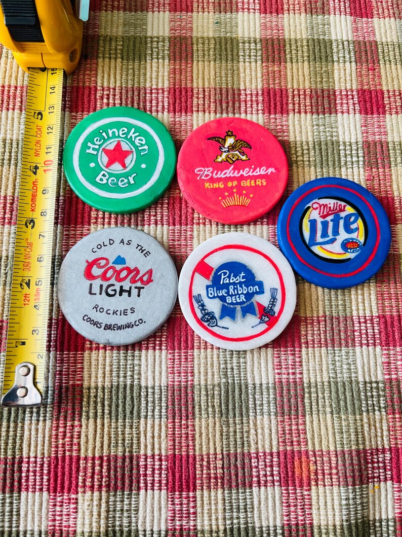 Puede incluir: Cinco fichas de cerveza redondas y de colores con diferentes logotipos de marcas. Las fichas son verde, roja, blanca, azul y morada. Los logotipos incluyen Heineken, Budweiser, Coors Light, Pabst Blue Ribbon y Miller Lite.