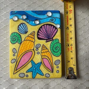 May include: A rectangular, hand-painted tile featuring a beach scene. The design includes a yellow sandy area, blue waves, and various seashells in orange, pink, and green. A blue starfish and clear, round embellishments are also present. The tile measures approximately 4 inches in length.