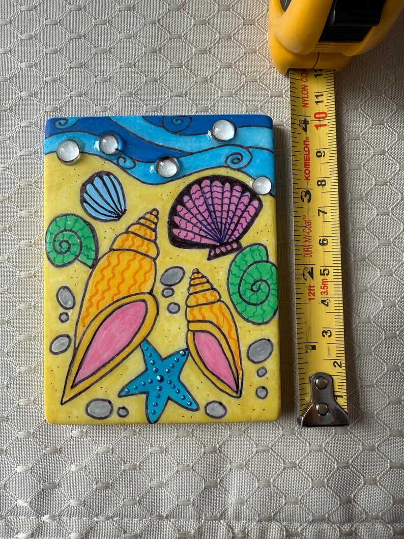 May include: A rectangular, hand-painted tile featuring a beach scene. The design includes a yellow sandy area, blue waves, and various seashells in orange, pink, and green. A blue starfish and clear, round embellishments are also present. The tile measures approximately 4 inches in length.