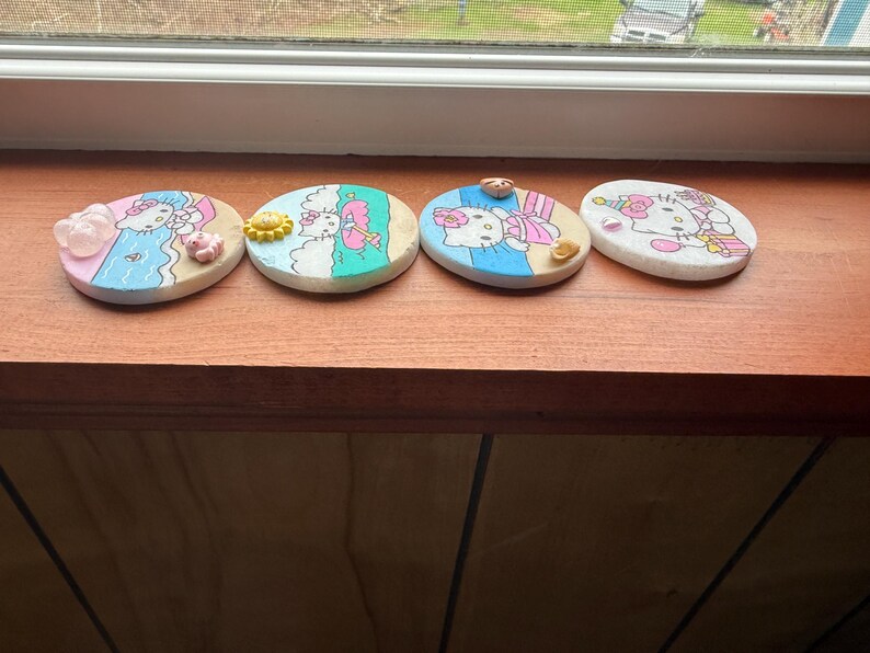 Op de afbeelding: Vier ronde onderzetters met een witte achtergrond en kleurrijke afbeeldingen van Hello Kitty op een strandthema. De onderzetters zijn versierd met kleine, kleurrijke versieringen.