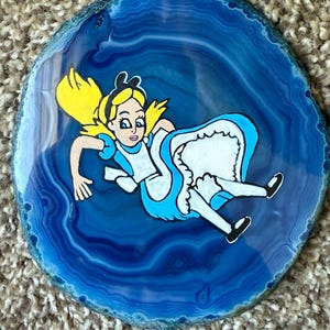 May include: A hand-painted blue agate slice featuring a cartoon illustration of Alice from Alice in Wonderland. Alice is depicted with blonde hair, a blue dress, and a white apron. The agate has a polished surface and a natural, organic shape.
