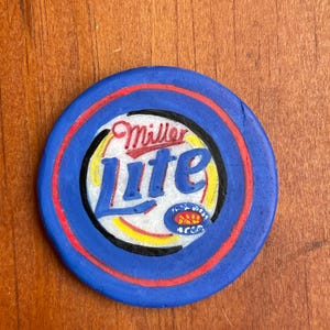 Puede incluir: Una ficha circular azul y roja con las palabras "Miller Lite" en letras blancas y negras. La ficha tiene un dise&ntilde;o amarillo y rojo en el centro.