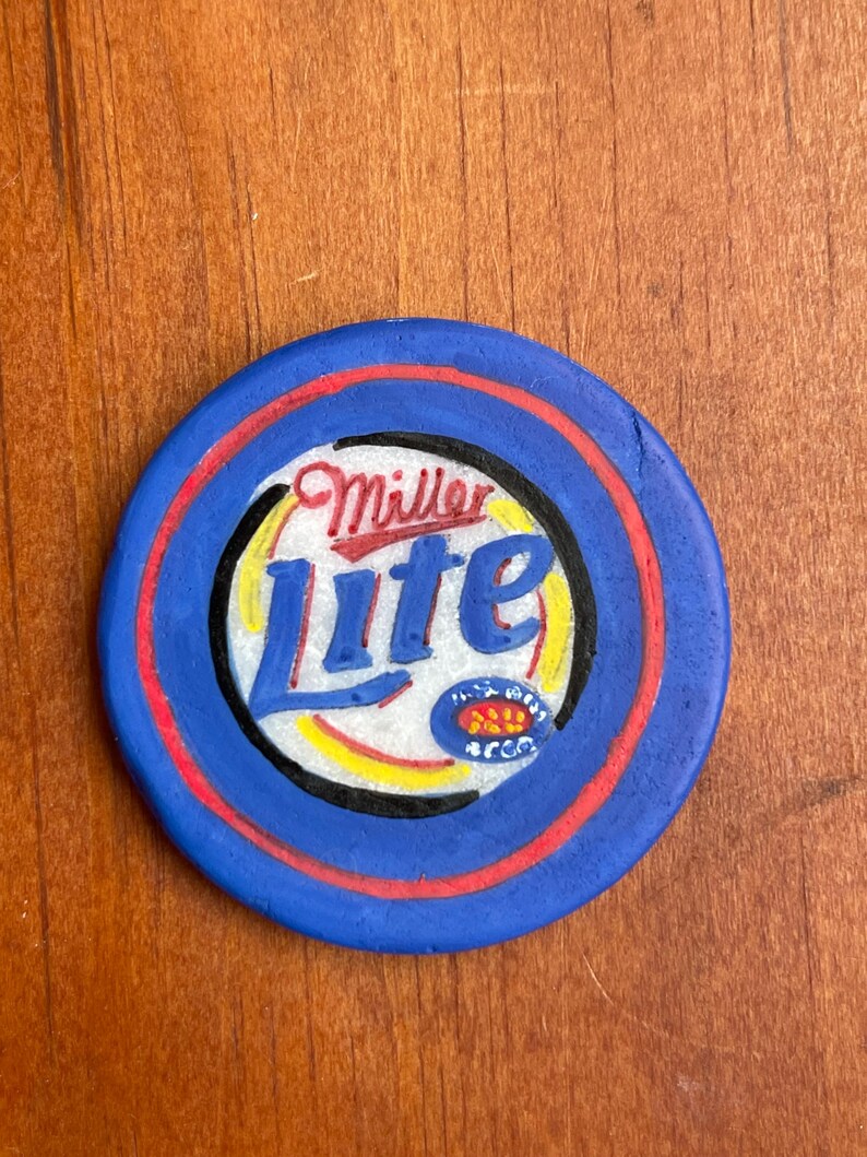 Puede incluir: Una ficha circular azul y roja con las palabras "Miller Lite" en letras blancas y negras. La ficha tiene un dise&ntilde;o amarillo y rojo en el centro.