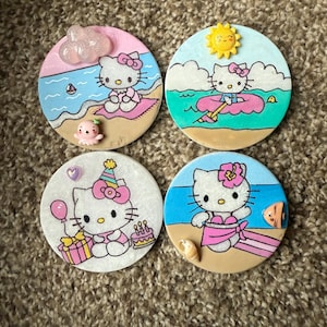Op de afbeelding: Vier ronde onderzetters met Hello Kitty in verschillende zomerse taferelen. De onderzetters zijn wit met een lichtroze, blauwe of gele achtergrond. Hello Kitty draagt een badpak, een zwemvest, een verjaardagshoed en een roze jurk. De onderzetters zijn versierd met schelpen, een zeilboot, een zon en een verjaardagstaart.
