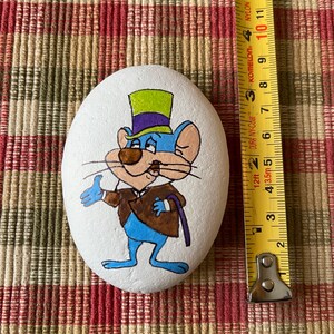 Puede incluir: Piedra pintada a mano con un rat&oacute;n de dibujos animados que lleva un sombrero de copa verde y morado y una chaqueta marr&oacute;n. El rat&oacute;n es azul con la nariz marr&oacute;n y los ojos blancos. La piedra est&aacute; pintada sobre un fondo blanco.