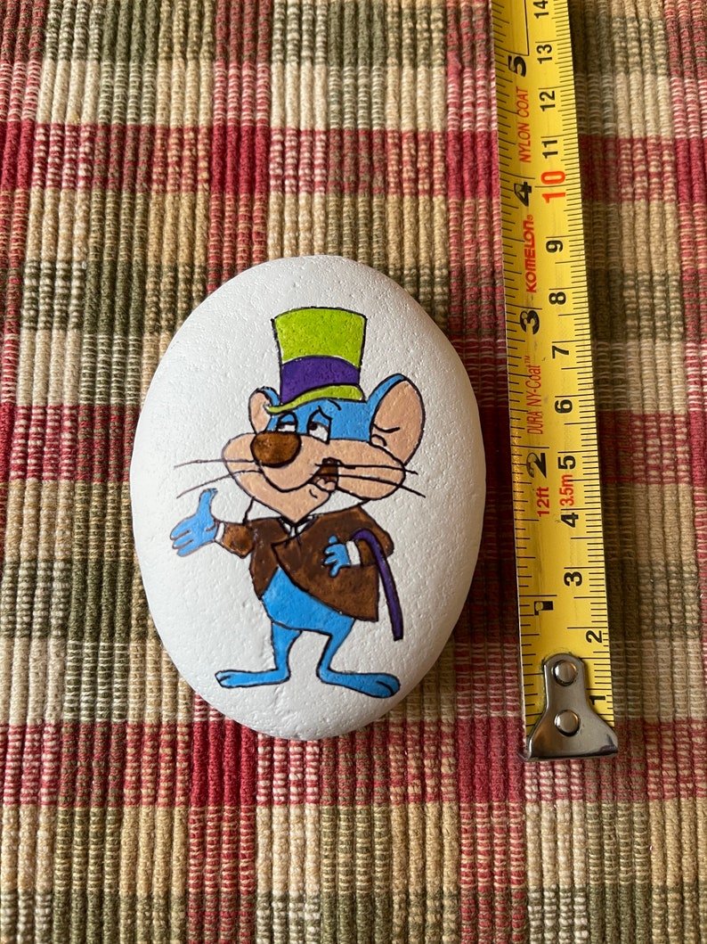 Puede incluir: Piedra pintada a mano con un rat&oacute;n de dibujos animados que lleva un sombrero de copa verde y morado y una chaqueta marr&oacute;n. El rat&oacute;n es azul con la nariz marr&oacute;n y los ojos blancos. La piedra est&aacute; pintada sobre un fondo blanco.
