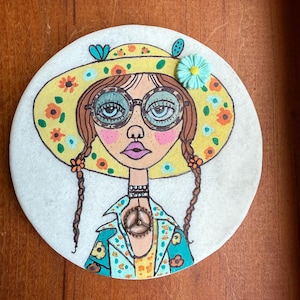 Puede incluir: Una piedra redonda pintada a mano con una ilustraci&oacute;n caprichosa de una mujer. Lleva un sombrero amarillo con flores, gafas y una chaqueta floral con un colgante de la paz. Su pelo est&aacute; trenzado. Tema bohemio.