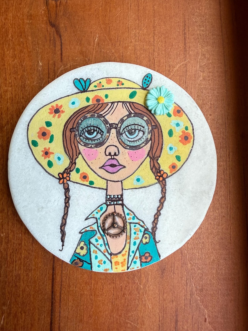 Puede incluir: Una piedra redonda pintada a mano con una ilustraci&oacute;n caprichosa de una mujer. Lleva un sombrero amarillo con flores, gafas y una chaqueta floral con un colgante de la paz. Su pelo est&aacute; trenzado. Tema bohemio.