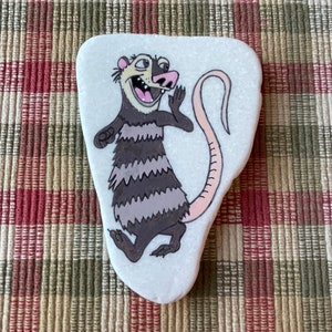 May include: Hand-painted rock art featuring a cartoon opossum with a striped tail and a surprised expression. The opossum is painted in shades of brown, gray, and pink on a white background.