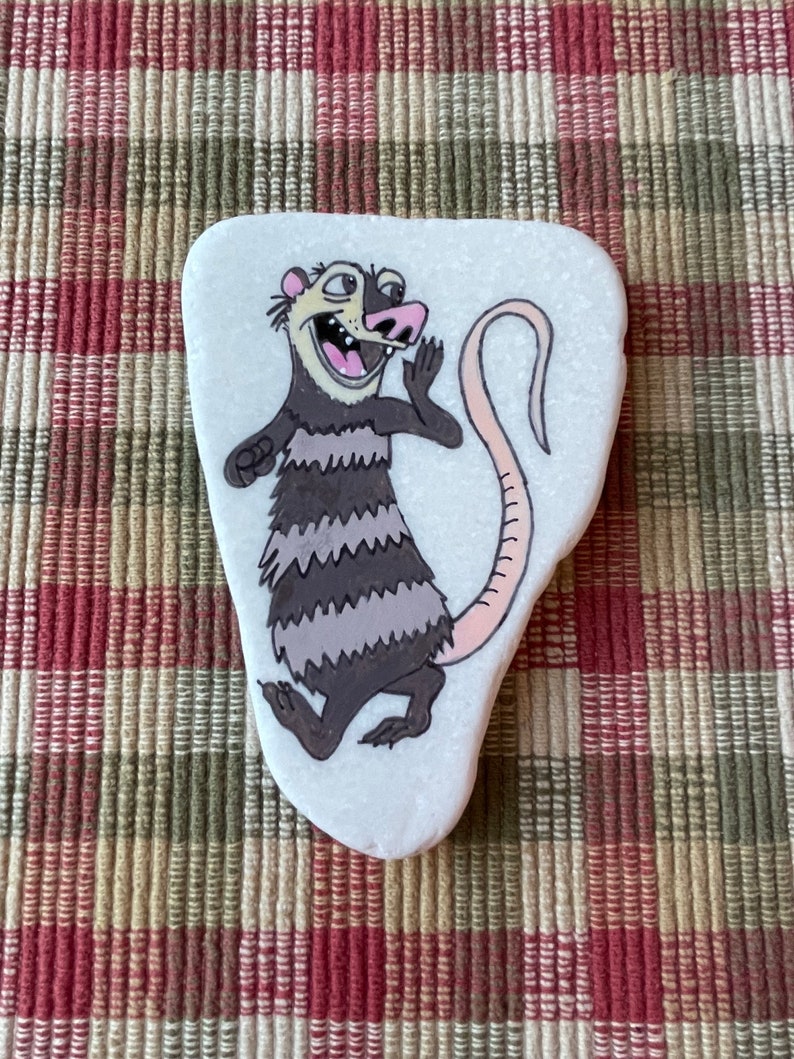 May include: Hand-painted rock art featuring a cartoon opossum with a striped tail and a surprised expression. The opossum is painted in shades of brown, gray, and pink on a white background.