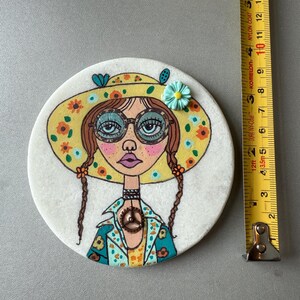 Puede incluir: Un posavasos redondo y blanco con una ilustraci&oacute;n pintada a mano de una mujer con un sombrero floral amarillo y gafas. El posavasos mide aproximadamente 10 cm de di&aacute;metro. La mujer tiene trenzas marrones y un colgante con el s&iacute;mbolo de la paz.