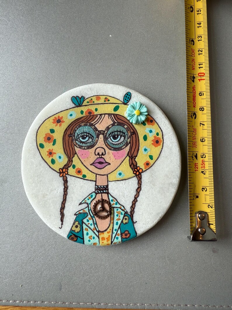 Puede incluir: Un posavasos redondo y blanco con una ilustraci&oacute;n pintada a mano de una mujer con un sombrero floral amarillo y gafas. El posavasos mide aproximadamente 10 cm de di&aacute;metro. La mujer tiene trenzas marrones y un colgante con el s&iacute;mbolo de la paz.