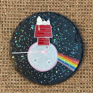 May include: A circular black slate coaster featuring a painted silver moon with a red doghouse on top. A white cartoon dog lies on the roof. A pink paintbrush extends from the moon, creating a vibrant rainbow trail against a glittery night sky.