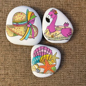 May include: Three hand-painted rocks with beach-themed designs. One rock features a yellow sun hat with a colorful bow. Another depicts a pink flamingo in a float. The third rock shows seashells and an orange starfish.