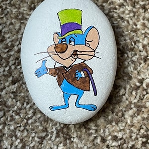 Puede incluir: Piedra pintada a mano con un rat&oacute;n de dibujos animados que lleva un sombrero de copa verde y morado y una chaqueta marr&oacute;n. El rat&oacute;n es azul con la cara blanca y bigotes negros.