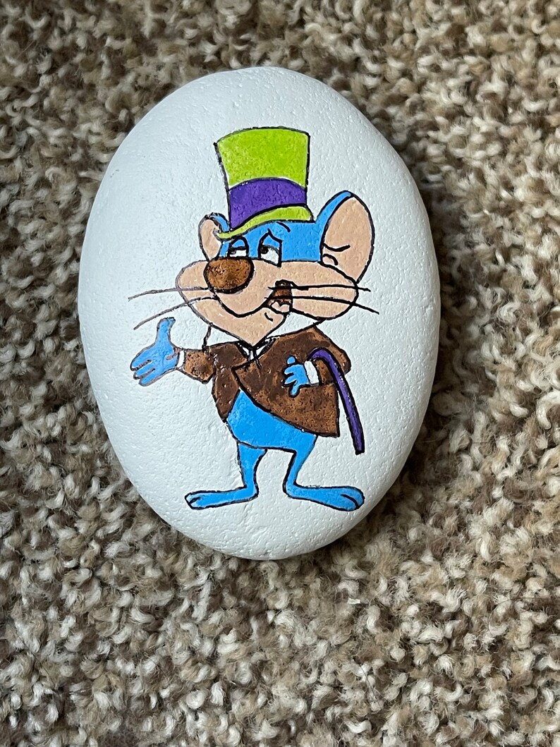 Puede incluir: Piedra pintada a mano con un rat&oacute;n de dibujos animados que lleva un sombrero de copa verde y morado y una chaqueta marr&oacute;n. El rat&oacute;n es azul con la cara blanca y bigotes negros.
