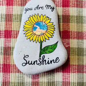 Puede incluir: Piedra pintada con un dise&ntilde;o de girasol. El girasol tiene un centro azul y amarillo y las palabras "You are my Sunshine" est&aacute;n escritas debajo de la flor.