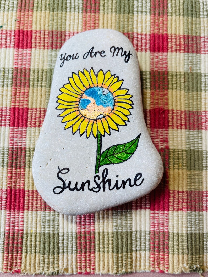 Puede incluir: Piedra pintada con un dise&ntilde;o de girasol. El girasol tiene un centro azul y amarillo y las palabras "You are my Sunshine" est&aacute;n escritas debajo de la flor.