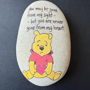 May include: A painted stone featuring Winnie the Pooh holding a pink heart. The text reads, "You may be gone from my sight - but you are never gone from my heart." The stone is oval-shaped and has a light beige color.